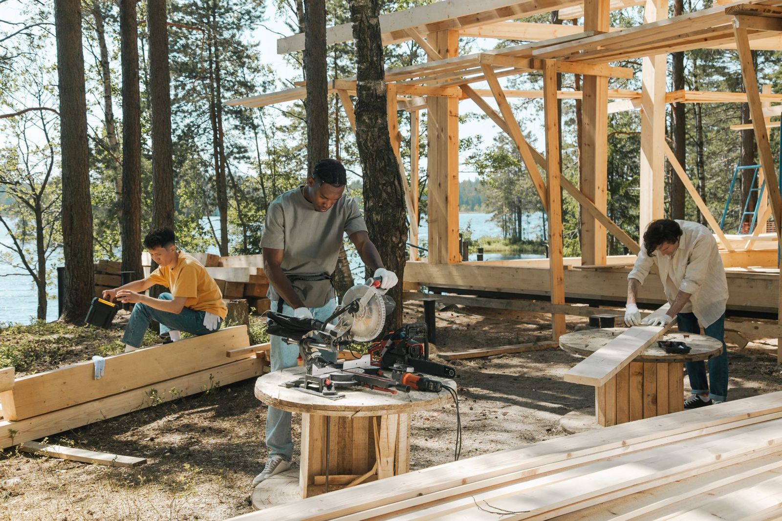 Three carpenters cutting and measuring lumber at an outdoor build site among pine trees next to a timber frame