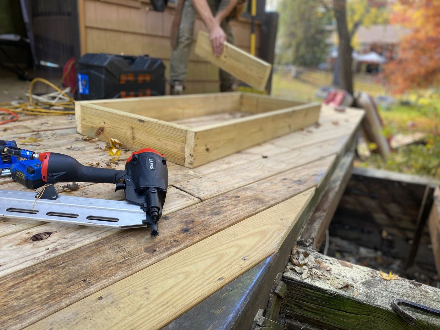 Framing nailer resting on a deck being resurfaced, with new pale boards beside weathered old ones and a worker carrying a board in the background