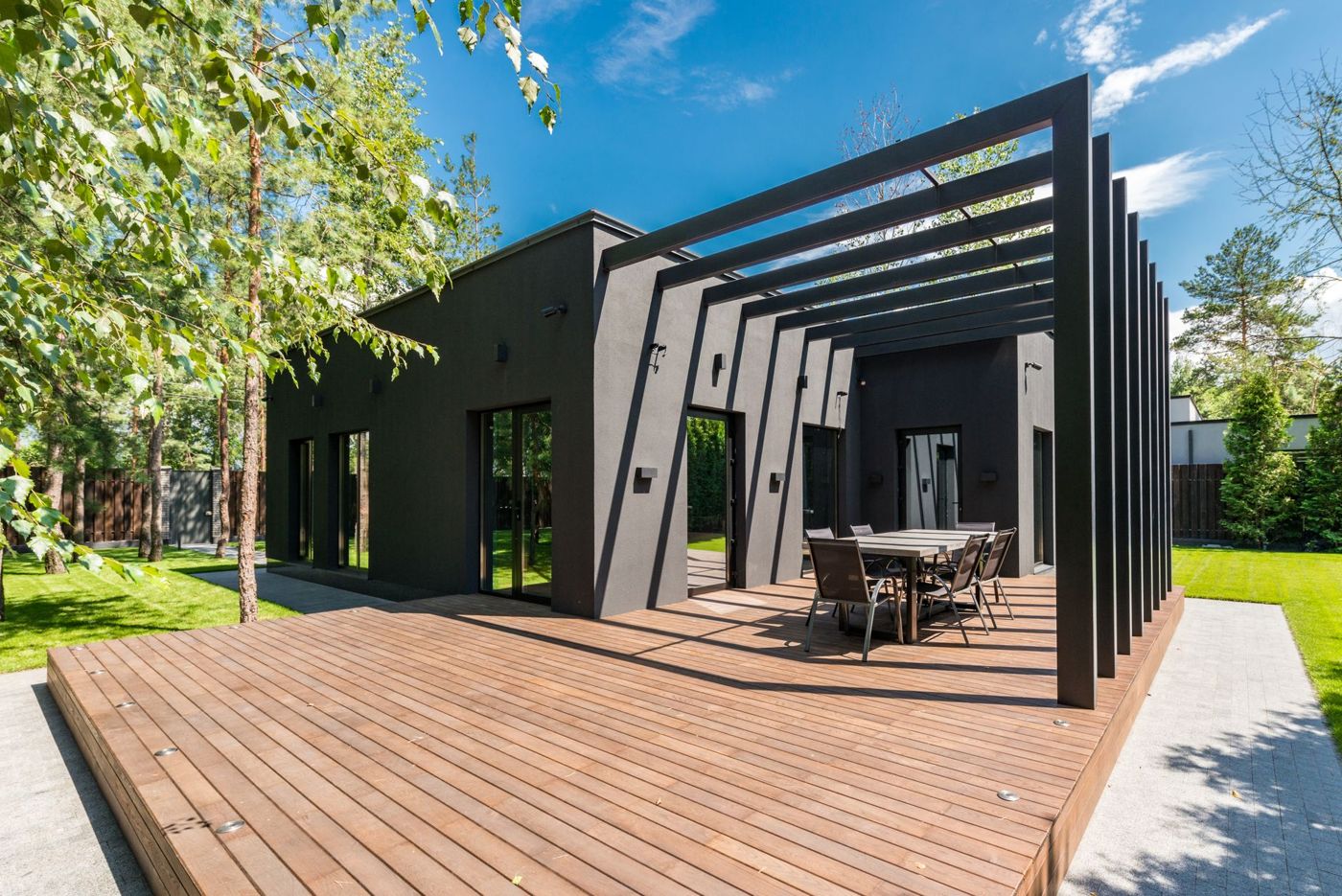 Large low-profile brown plank deck with small built-in lights, an outdoor dining table and a black slatted pergola beside a dark modern house surrounded by trees