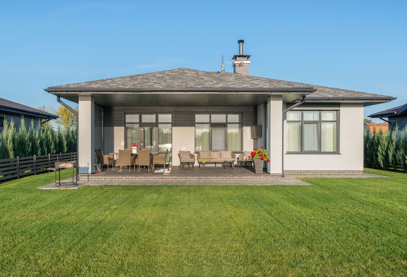 Single-story gray house with a covered back patio holding wicker lounge and dining furniture beside a wide lawn and a dark horizontal fence