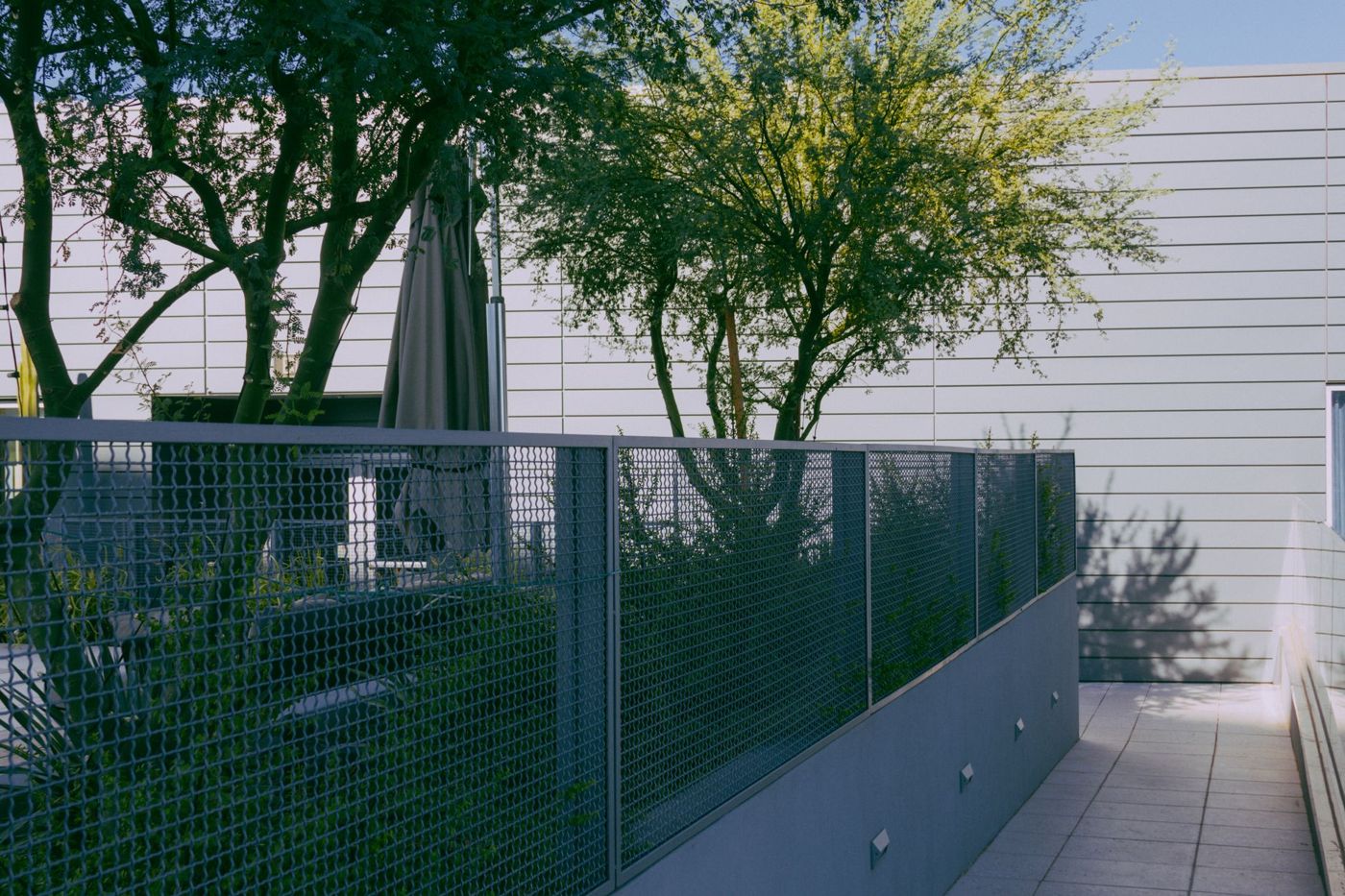 Framed galvanized mesh fence panels on a low gray wall along a paved walkway with trees behind