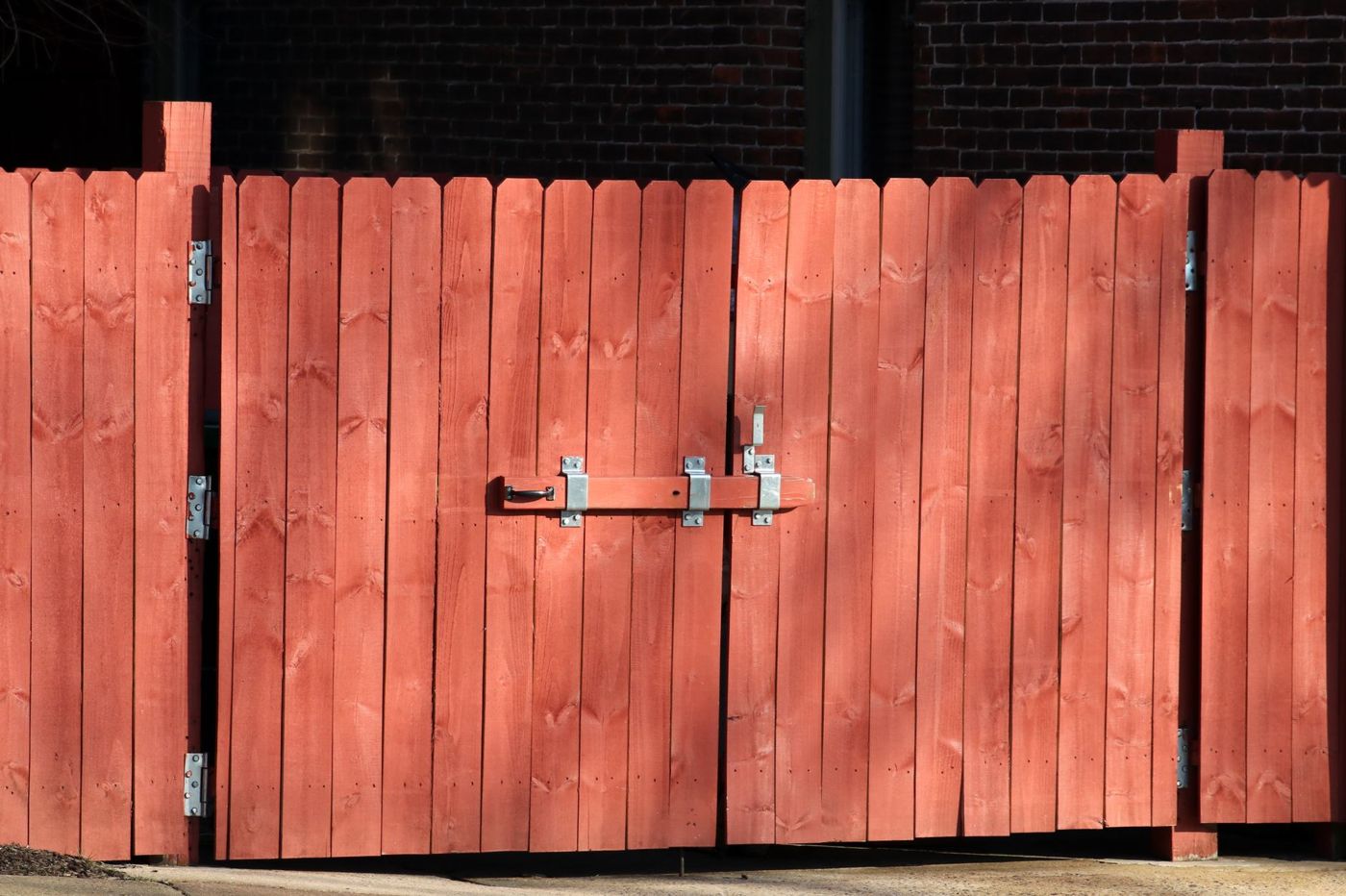 Red-stained double wooden gate in a dog-ear board fence with galvanized strap hinges and a wooden drop-bar latch across both leaves