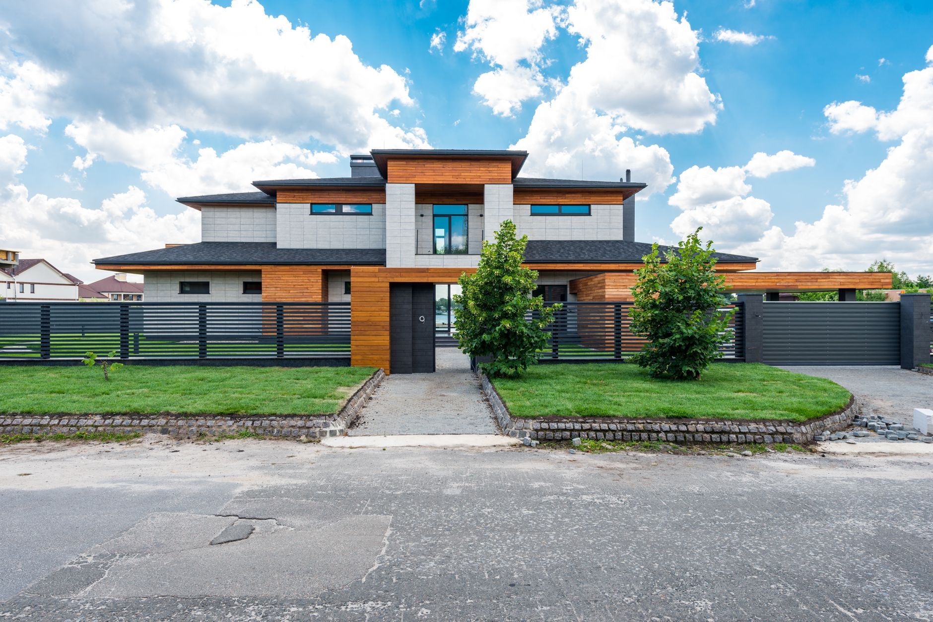 Modern two-story house behind a dark horizontal-slat metal fence with a wood-clad entry wall, a matching sliding gate and a paver path across a fresh lawn