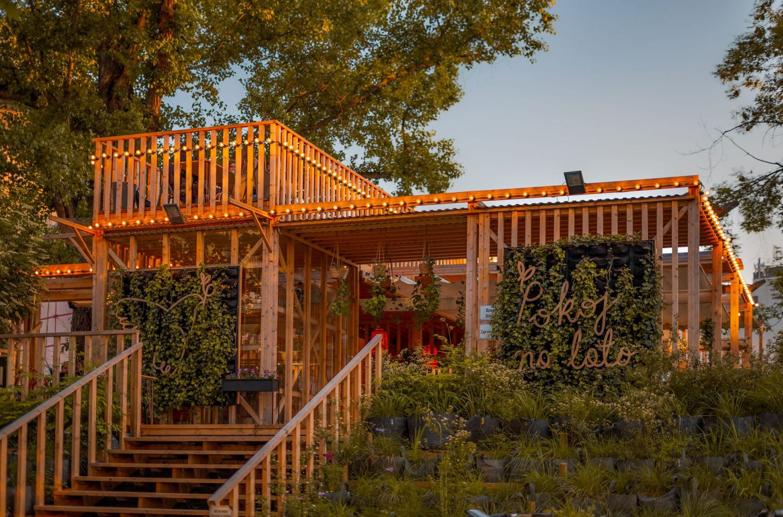 Wooden stairs with slatted railings climbing to a raised timber pavilion with string lights and a planted hillside at sunset