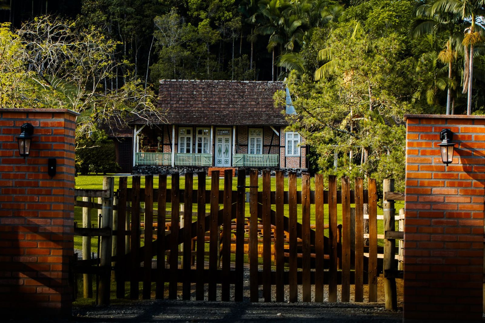 Double wooden picket gate between red brick pillars with lanterns, opening onto a lawn and a small house among trees